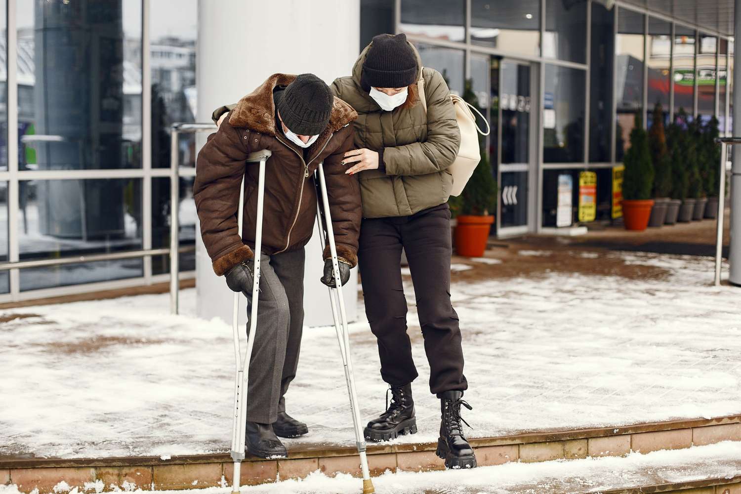 A person using crutches is helped by another individual while walking on a snowy, icy sidewalk outside a building, illustrating winter slip and fall risk.