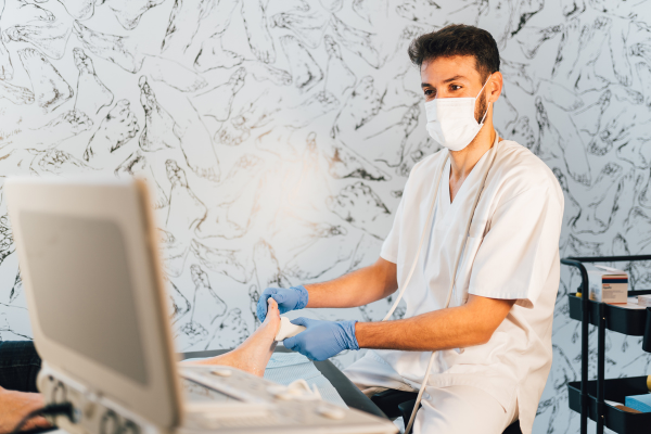 Podiatrist in blue gloves and face mask performing an ultrasound-guided examination of a patient's foot with imaging equipment, used for gout diagnosis and intra-articular injection guidance at Foot & Ankle Centers of New Jersey